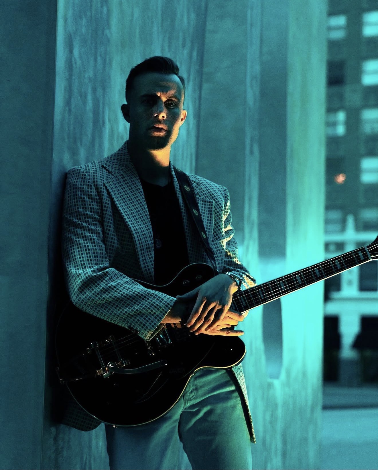 Tom Lail in a checkered blazer holding a black electric guitar in blue evening light